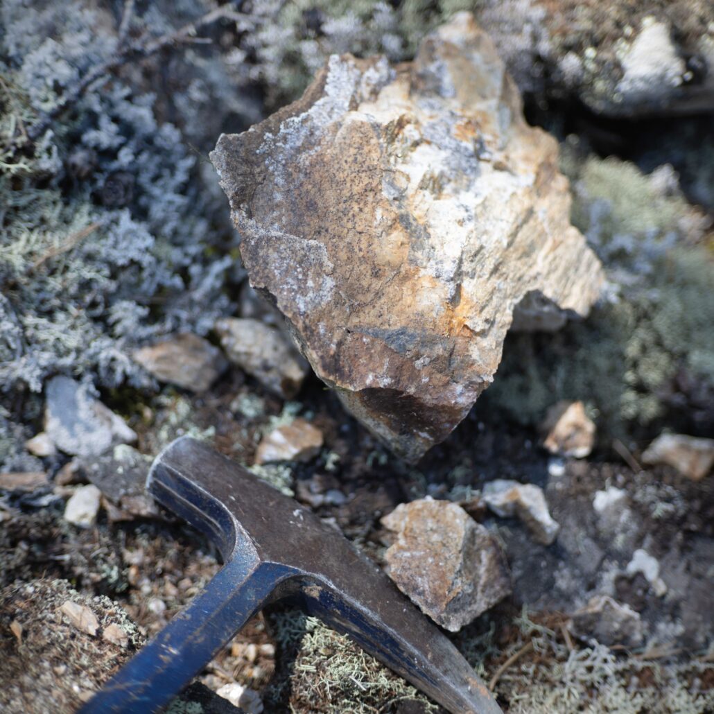Rock sample beside a geological hammer during field prospecting for mineral exploration.